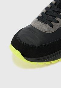 Zwarte en grijze sneaker met een gestructureerde bovenkant van suede en synthetische materialen, voorzien van een felgele rubberen zool en zwarte veters.