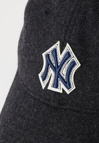 Grå baseballcap i en blød struktur med en hævet broderet blå og hvid "NY" logo. Har en buet skygge og tekstureret stof.