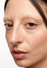 Nahaufnahme eines Gesichts mit glatter, heller Haut, geradem Nasenprofil, vollen Lippen mit natürlichem Glanz und braunen Augen. Die Augenbrauen sind hell und fein.