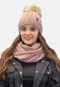 Bonnet en tricot rose avec pompon, motif texturé ; associé à une écharpe tricotée rose assortie. Porté sur une veste noire. Accents argentés visibles.