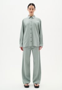 Lichtgroen linnen shirt en wijde broek met een knopenlijst aan de voorkant. Heeft een kraag, lange mouwen en een relaxte pasvorm. Schoenen zijn zwarte platte sandalen.