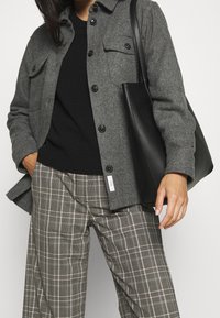 Grå uld skjortejakke med knaplukning, sort sweater under, ternede bukser i grå og hvid, og en sort læder tote bag.