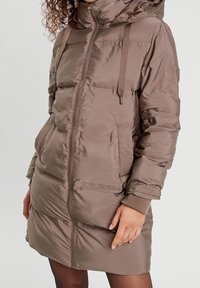 Abrigo acolchado marrón con cuello alto, cremallera frontal, dos bolsillos laterales y puños acanalados. Textura suave y brillante con un diseño de longitud media.
