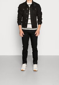 Chaqueta vaquera negra con detalles desgastados, camiseta blanca con rayas debajo, pantalones negros y zapatillas blancas con detalles en naranja.