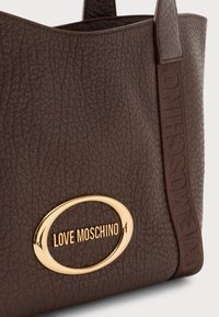 Tuvuma skats uz tekstūrētu brūnu ādas tote somu ar zelta ovālo "Love Moschino" logo un atbilstošu brūnu siksniņu ar iespiestu zīmola nosaukumu.