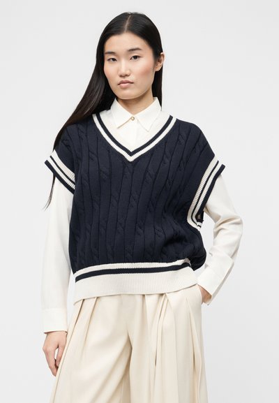 Femme aux longs cheveux noirs portant un gilet sans manches en tricot torsadé bleu marine sur une chemise blanche à col et un pantalon large crème.