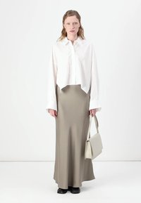 Weißes, kurzes Button-Up-Shirt, khakifarbener Satin-Maxi-Rock, schwarze Schuhe und eine hellgraue Handtasche, vor einem neutralen Hintergrund stehend.