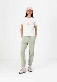 Witte katoenen t-shirt met groene en rode grafische print, gecombineerd met lichtgroene joggers en een roze pet. Sneakers zijn lichtgrijs met donkere accenten.