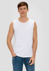 Young man standing with hands partially in pockets, wearing a white sleeveless shirt and blue jeans against a plain background.