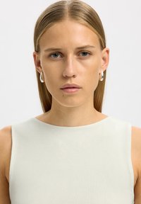 Jeune femme aux cheveux blonds et lisses, portant des boucles d'oreilles argentées en anneau et un haut blanc sans manches, regardant directement la caméra.