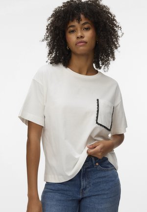 Femme aux cheveux bouclés portant une chemise blanche à manches courtes avec une poche bordée de noir et un jean bleu, debout devant un fond uni.