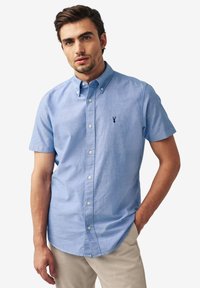 Šviesiai mėlynas trumpomis rankovėmis marškinėliai su mygtukais, pagaminti iš minkštos medžiagos, su klasikiniais apykaklėmis ir tamsiai mėlyna logotipo siuvinėjimu krūtinėje.