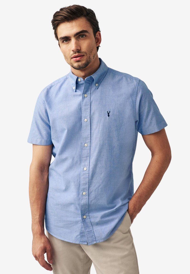 Šviesiai mėlynas trumpomis rankovėmis marškinėliai su mygtukais, pagaminti iš minkštos medžiagos, su klasikiniais apykaklėmis ir tamsiai mėlyna logotipo siuvinėjimu krūtinėje.