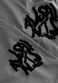 Tela de algodón gris con un destacado logo de terciopelo negro que dice "ALLSAINTS LONDON" en una tipografía estilizada y abstracta.