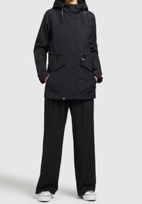 Musta vedenpitävä takki hupulla, säädettävillä kiristysnarulla, kahdella etutaskulla ja napillukolla, yhdistettynä löysiin mustiin housuihin ja valkoisiin tennareihin.