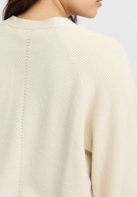 Ljust beige randig tröja med rundad halsringning och subtila sömdetaljer baktill, som visar en texturerad stickad design och avslappnad passform.
