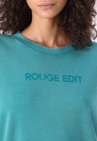 Türkiissinine T-särk, millel on ümar kaelus ja lühikesed varrukad. Ees on tekst "ROUGE EDIT", mis on veidi tumedamas varjus.