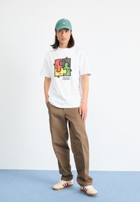 Weißes T-Shirt mit einem bunten Puzzlestück-Design. Braune Hose, beige Schuhe mit roten Streifen. Helle bläuliche Bodenhintergrund. Grünes Cap.