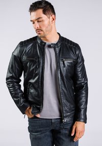 Chaqueta de cuero negra con detalle acolchado en los hombros, bolsillos con cremallera y cuello alzado, llevada sobre una camisa gris y vaqueros azules.