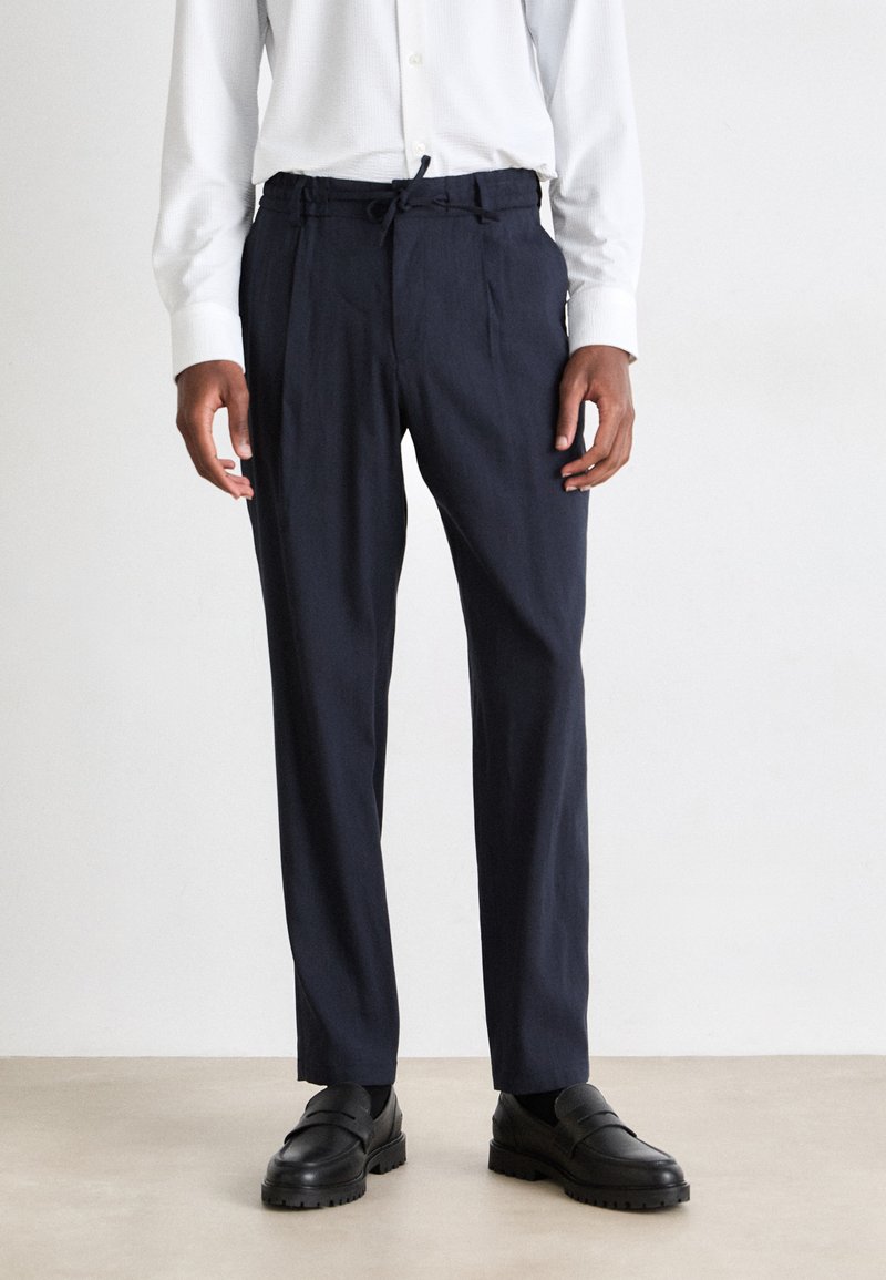 Pantalones entallados azul marino con cintura elástica, fabricados en tejido ligero, con pliegues frontales y bolsillos laterales, combinados con mocasines negros.