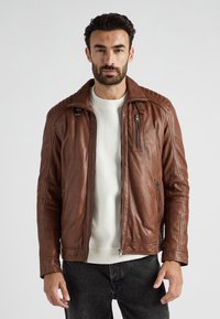 Bruine leren jas met een rits aan de voorkant, gewatteerde schouderaccenten en zijzakken, gedragen over een licht truitje en donkere spijkerbroek.