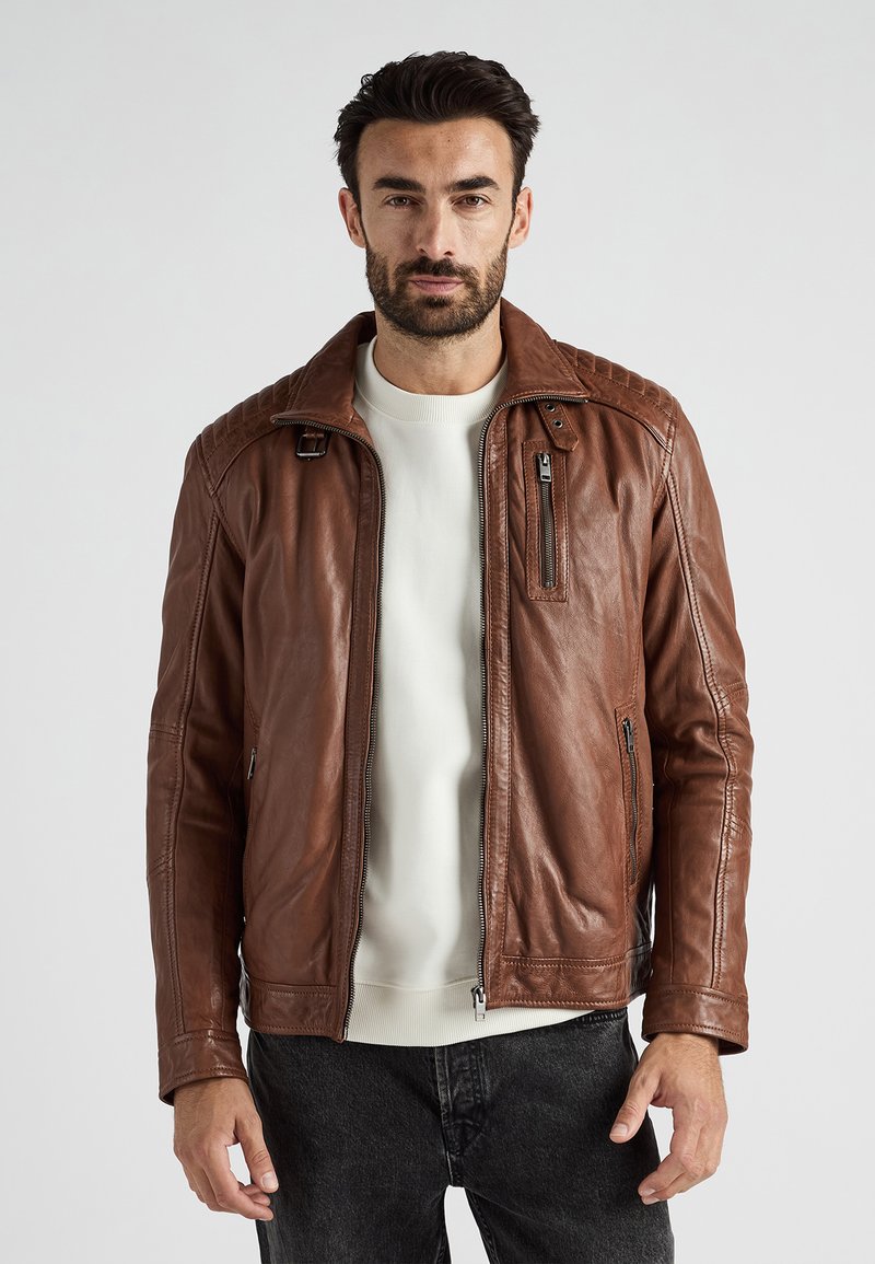 Bruine leren jas met een rits aan de voorkant, gewatteerde schouderaccenten en zijzakken, gedragen over een licht truitje en donkere spijkerbroek.