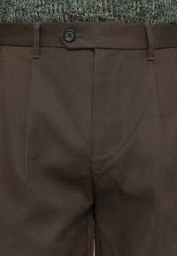 Brown trousers with side pockets, a buttoned waist tab, belt loops, and a multicolored knitted sweater tucked in.