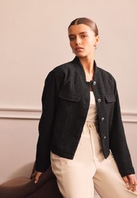 Giacca nera con texture, scollo tondo, tasche frontali e bottoni argentati, abbinata a pantaloni chiari. Design semplice e moderno.