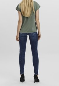 Žena s dlouhými blond vlasy, oblečená v zeleném tričku, tmavě modrých skinny džínách a černých vysokých podpatcích, stojící zády.