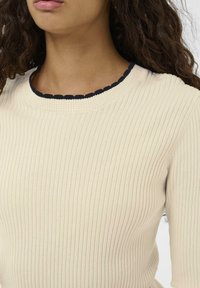 Kvinde iført en cremefarvet ribstrikket sweater med en sort bølget kant ved halsudskæringen, med krøllet, mørkt hår synligt over skulderen.