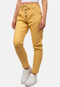 Pantalon en coton jaune avec une coupe slim, doté d'une taille à cordon de serrage et de quatre boutons. Porté avec des baskets blanches.