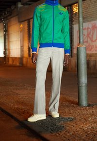 Vihreä ja sininen vetoketjullinen takki raidallisilla yksityiskohdilla, yhdistetty vaaleanharmaisiin ruudullisiin housuihin. Asun täydistää valkoiset tennarit.