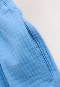 Shorts en tissu bleu clair avec taille élastique et motif carré texturé, montrant la couture latérale et une partie de l'ouverture de la poche.