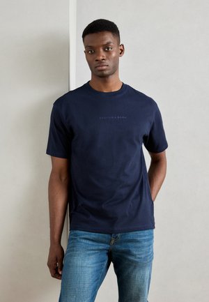 Navy blue short-sleeve t-shirt features a crew neck and the text "SCOTCH & SODA" printed subtly on the chest. Worn with light blue jeans.