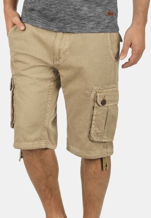 Beige cargoshorts med sidelommer båret af en person, der står, kombineret med en grå meleret skjorte.