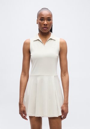 Φωτεινό μπεζ φόρεμα χωρίς μανίκια με γιακά πόλο, φουσκωτή φούστα και λείο υλικό. Διαθέτει μινιμαλιστικό σχέδιο με διακριτικό λογότυπο στο στήθος.