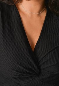 Top avvolgente nero con texture, caratterizzato da un profondo scollo a V, dettaglio arricciato al centro e un sottile luccichio. Accentuato da una sottile collana choker d'argento.