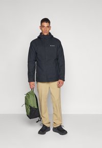 Veste noire imperméable avec capuche, portée par-dessus un pantalon beige. La personne tient un sac à dos vert et noir, portant des chaussures noires.