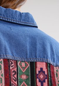 Giacca in denim con un pannello in tessuto intrecciato colorato sulla schiena, caratterizzato da motivi geometrici in rosso, verde e nero, e un design del colletto classico.
