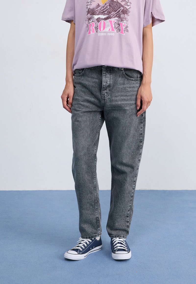 Vaqueros de mezclilla gris con un corte relajado, que cuentan con cinco bolsillos y un cierre de botón, combinados con una camiseta gráfica de color lila claro y zapatillas azules.