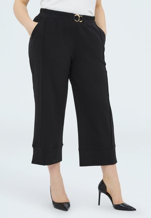 Femme portant un pantalon court noir taille haute avec une ceinture noire à double boucle dorée et des escarpins noirs à bout pointu, les mains dans les poches.