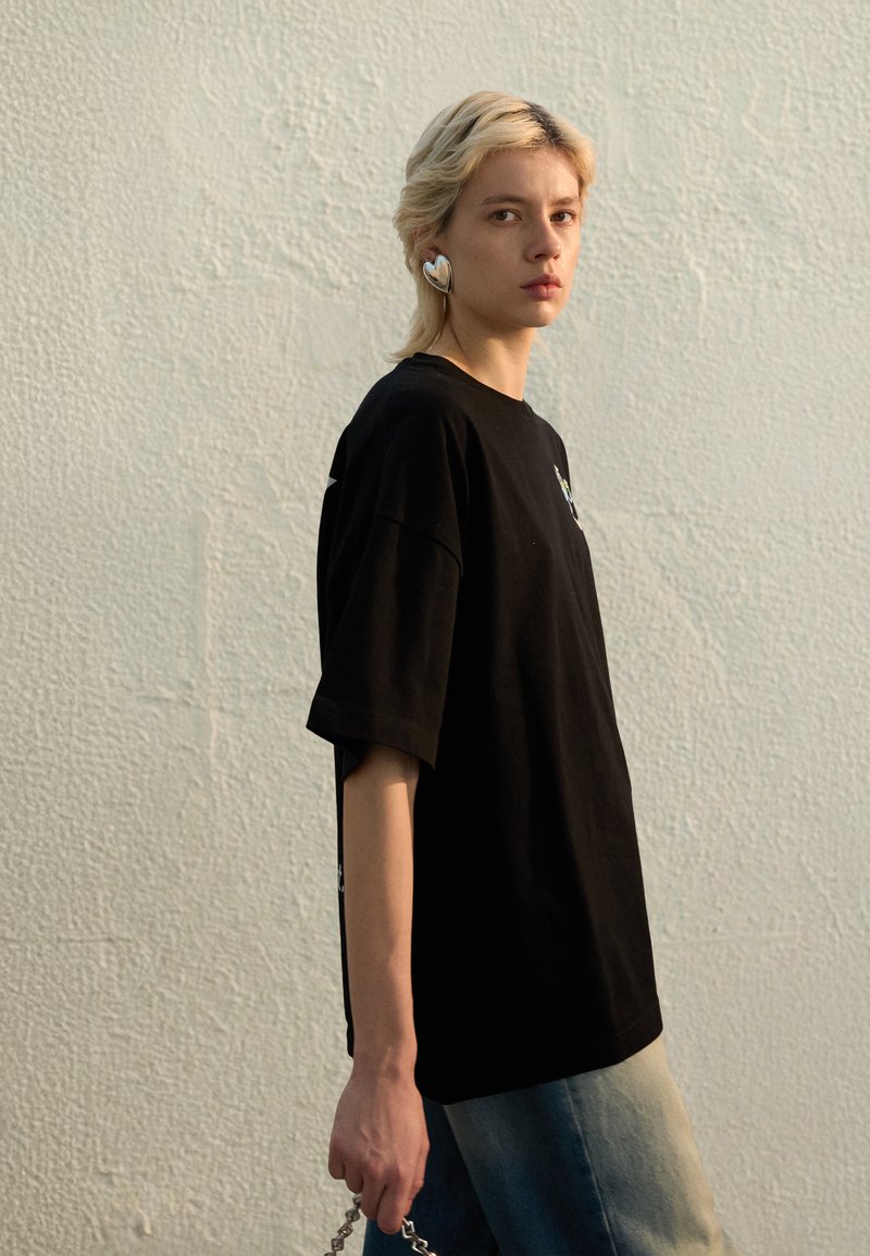 Black oversized t-shirt with short sleeves, rounded neckline, and graphic detail. Paired with light blue denim and a silver chain accessory.