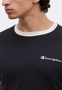 Άνδρας που φοράει μαύρο T-shirt με λευκό γιακά, λευκό κεντημένο λογότυπο "Champion" στο αριστερό στήθος, στραμμένος προς τα αριστερά, κομμένο στον λαιμό και τον ώμο.