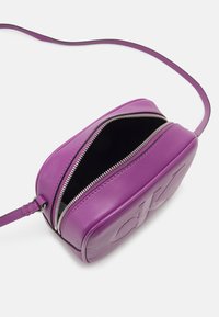 Borsa a tracolla in pelle viola con zip argentata parzialmente aperta che mostra l'interno nero, e lunga tracolla regolabile su sfondo bianco.