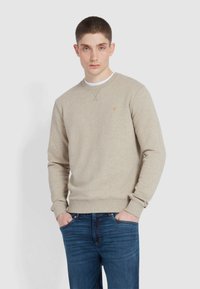 Beżowy sweatshirt z okrągłym dekoltem, ściągaczami przy rękawach i u dołu. Posiada małe pomarańczowe logo na klatce piersiowej. W zestawie z ciemnoniebieskimi dżinsami.