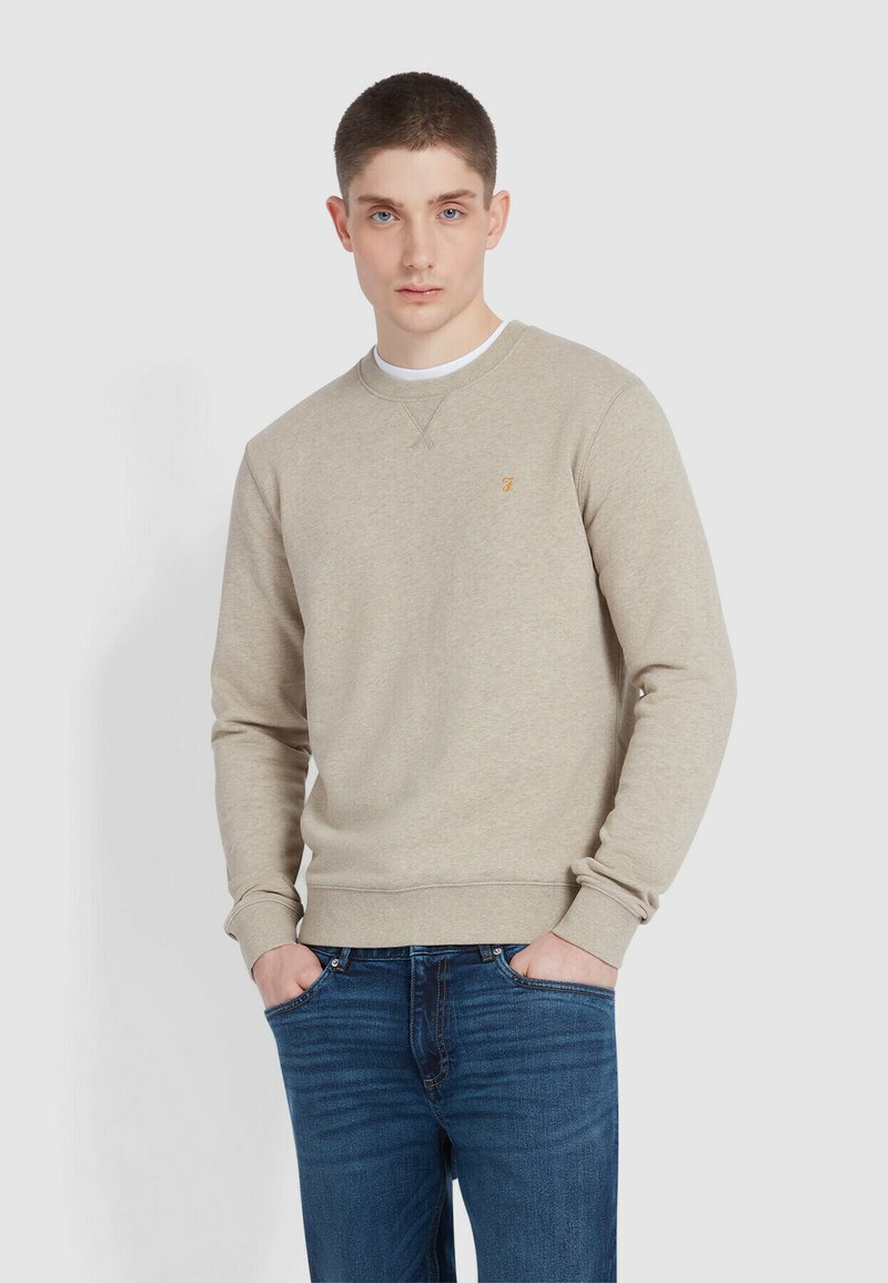 Beżowy sweatshirt z okrągłym dekoltem, ściągaczami przy rękawach i u dołu. Posiada małe pomarańczowe logo na klatce piersiowej. W zestawie z ciemnoniebieskimi dżinsami.