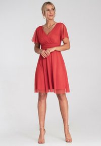 Rotes knielanges Kleid mit kurzen Schmetterlingsärmeln, einem V-Ausschnitt und einem taillierten Oberteil. Der durchsichtige Überwurf und der fließende Rock erzeugen Bewegung.