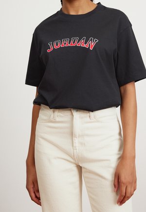 Osoba ubrana w czarną koszulkę z nadrukiem "JORDAN" w czerwonych i białych literach, zestawioną z wysokim stanem jasnobeżowych spodni.