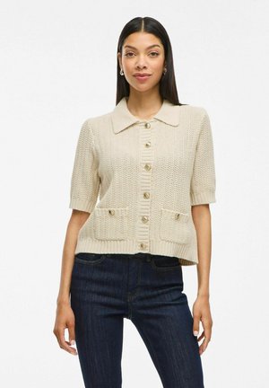 Cardigan à manches courtes, couleur crème, avec un col, un devant boutonné et deux poches à l'avant. Tissu en tricot texturé.