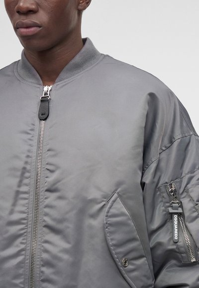 Gros plan d'une personne portant une veste bomber grise avec fermeture éclair frontale, poche à rabat et poche zippée sur la manche avec une étiquette noire.